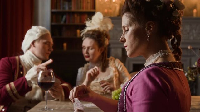 Side medium close-up of mature lavishly dressed Victorian noblewoman sitting at dinner table, holding hand of cards, while lord and lady friends are whispering, discussing game strategy