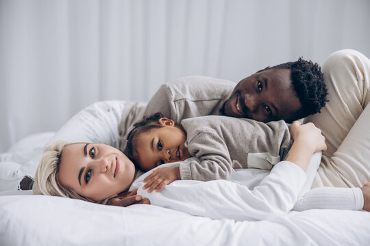 Happy african man and caucasian woman resting on bed with their biracial toddler daughter. - Powered by Adobe
