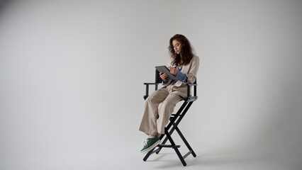 Fashionable Model Sitting on Director's Chair with Tablet in Studio Setting