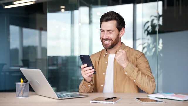 Satisfied happy handsome businessman received great news on mobile phone while sitting at workplace in business office. Smiling glad bearded entrepreneur reads a positive good message on a smartphone