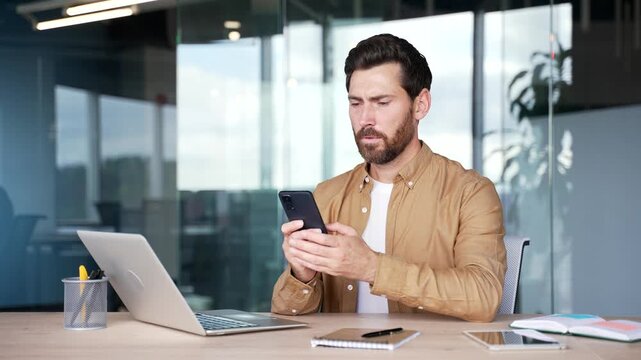 Upset worried asian businessman reads bad news on mobile phone while sitting at desk at workplace in modern business office. Frustrated worker is sad while reviewing negative information on smartphone