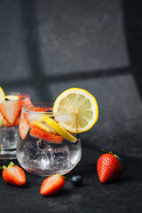 Sparkling lemonade with lemon slices strawberries and blueberries in glass on dark background with fresh fruits nearby