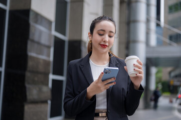 Fototapeta premium A smiling office woman is standing outside an office building, holding a cup of coffee while checking her smartphone.