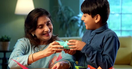 Indian grandfather and kid doing paper craft together at home, teaching colorful boat or aeroplane using bright papers, elderly man teaching grandson creative activity, family bonding indoors
