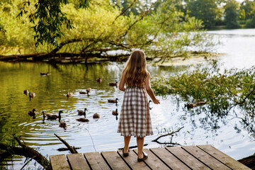 Little elementary school girl feeding wild ducks on a pond. Cute child enjoying nature and animals outdoors in summer, happy kid with eye glasses spending time near water.