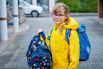 Obraz premium Happy little elementary school girl with eyeglasses and satchel backpack walking outdoors on the way to school. Smiling pupil child ready for back to school, education and childhood lifestyle concept.