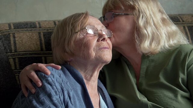 Tender moment elderly woman receiving loving kiss and embrace from adult daughter. Family warmth and affection on cozy couch at home. Care of old people. Meeting relatives of dementia patients