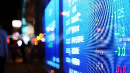 A close-up of a bright blue LED stock market ticker displays fluctuating financial data at night, with a bustling, out-of-focus city street behind it. - Powered by Adobe