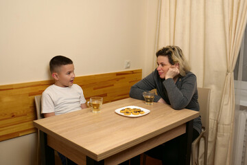 Mother and son sitting at table, drinking tea and talking with cookies on plate, sharing family time in warm cozy home.