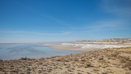 Expansive desert landscape with rugged terrain, white limestone cliffs, and a serene reflective water body under a clear blue sky, located in a remote arid region.