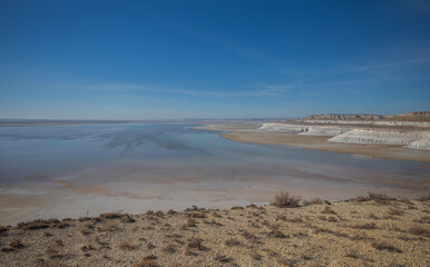 A vast salt plain with white cliffs and reflective water under a clear blue sky, surrounded by barren desert terrain. Located in Mangystau, Kazakhstan.