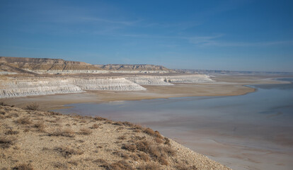 A vast salt plain with white cliffs and reflective water under a clear blue sky, surrounded by barren desert terrain. Located in Mangystau, Kazakhstan.