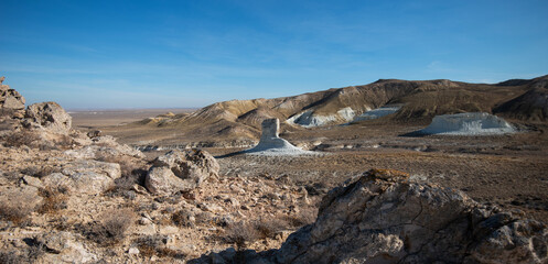A rugged desert landscape stretches across rolling hills, with a striking isolated rock formation standing in the valley below. The arid terrain under a clear blue sky showcases the natural beauty.