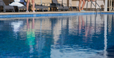 Reflection of people on the calm surface of a swimming pool in a sunny resort setting, with pool chairs and loungers in the background, creating a relaxing and peaceful atmosphere.