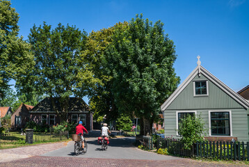 Two cyclists ride down a quiet street in a charming village with traditional houses and lush greenery, enjoying a peaceful summer day under a clear blue sky, surrounded by trees and gardens.