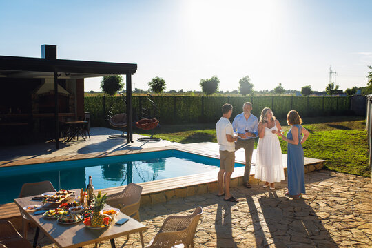 Group of adult friends talking by swimming pool drinking wine in sunny backyard. - Powered by Adobe