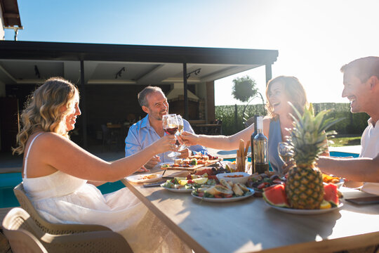 Group of adult friends having lunch by swimming pool in sunny backyard and toasting with wine.