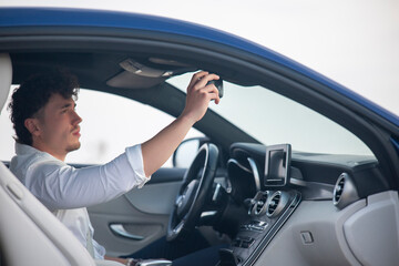 Young man inside luxury blue car adjusting rearview mirror with focused expression