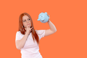 Woman contemplating finances while holding blue piggy bank against a bright orange background in a casual setting during the day