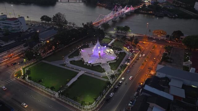 Night aerial of Surat Thani City Pillar Shrine glowing, with city lights, Tapi River, and traffic illuminating the urban landscape of Thailand.