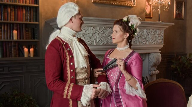 Medium shot of young Caucasian noblewoman in pink silk dress, holding fan, standing in manor parlour with courteous fiance in wig, lace cravat and red surcoat, chatting and flirting