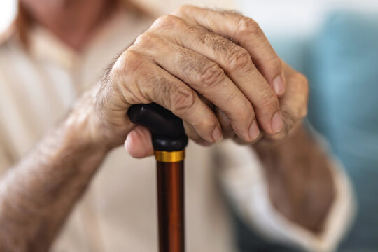 Senior man sitting on the sofa and looking contemplative while holding his walking stick. Walking difficulties. Serious bearded senior leaning on walking stick while thinking about his helplessness. - Powered by Adobe