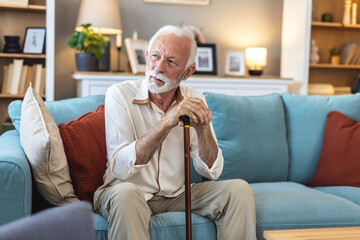 Senior man sitting on the sofa and looking contemplative while holding his walking stick. Walking...