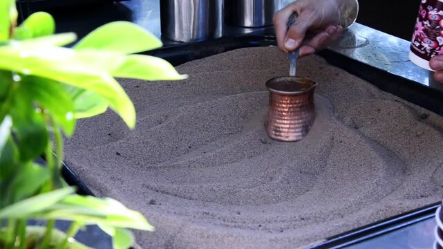 Close-up of a hand holding a cezve (copper coffee pot) brewing traditional Turkish coffee in hot sand.