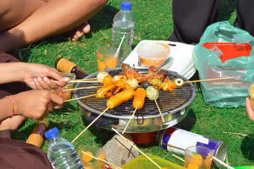 A detailed view of portable grill with skewered sausages, chicken pieces, condiments, and a plastic water bottle placed neatly on the grassy ground, capturing the essence of picnic cooking.