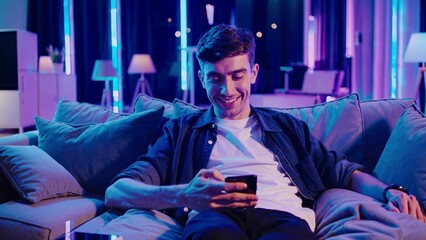 Obraz premium Man sitting on couch with his phone. Man relaxing on a smartphone. Modern casual room with a modern atmosphere. A man is resting on a sofa while using his phone lifestyle.