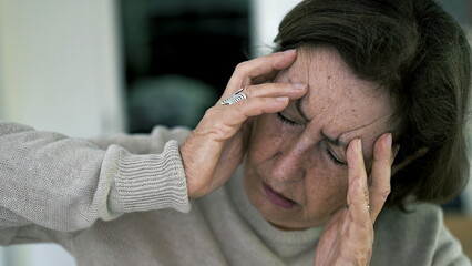 Senior woman in visible distress, holding head with eyes closed, fighting severe headache. Facial...