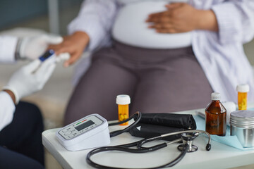 Pregnant Black woman sitting while healthcare professional measuring blood glucose level, medical equipment including blood pressure monitor and stethoscope on table in foreground