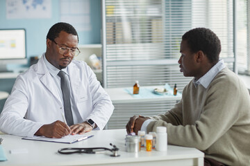 Black middle aged male doctor writing notes while consulting Black young adult man in medical office, prescription bottles and stethoscope on desk, both seated facing each other