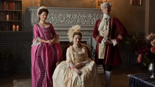 Medium full portrait of 18th century Caucasian gentleman, wearing red surcoat, breeches, wig and two glamorous ladies in glamorous silk dresses sitting in armchair and standing in mansion parlour