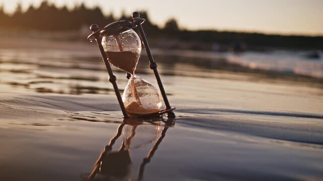 Glass Hourglass With Golden Sand Ticking Away On Coastline, Surrounded By Ocean Breeze And Sunset Glow, Representing Time, Tranquility, And Life Fleeting Moments.