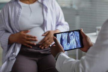 Pregnant Black woman sitting with hands on belly while medical professional holding tablet...