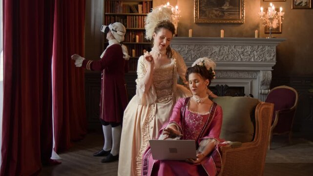 Full shot of two beautiful actresses in opulent Victorian costumes browsing online on laptop on 18th century movie set, while actor with VR headset is exploring historical context in background