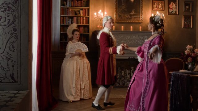Full shot of aristocratic 19th century couple, woman in pink dress and gentleman in red frock, breeches dancing together at ball in royal palace, while lady with fan is standing by and watching