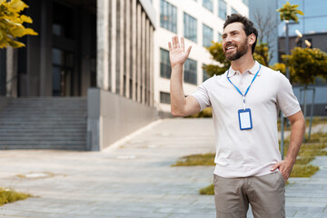 Smiling businessman waving hand, wearing badge, standing outside office building
