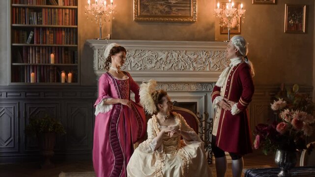 Medium full shot of two aristocratic ladies in glamorous dresses and man in historical frock coat having fun conversation in palace, gentleman entertaining female friends with stories