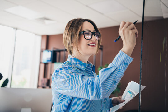 Confident businesswoman writing on a glass board in office workspace with documents, presenting ideas for successful project