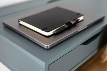 Black notebook with pen placed on closed silver laptop lying on wicker wooden table next to bed inside cozy modern home interior with soft natural light