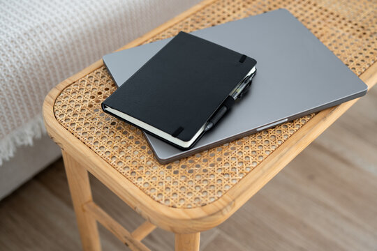 Black notebook with pen placed on closed silver laptop lying on wicker wooden table next to bed inside cozy modern home interior with soft natural light