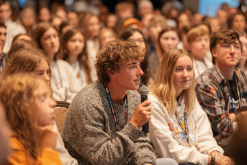 Young man asking a question into a microphone during a conference, with audience members listening attentively