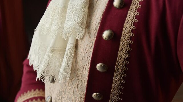 Close-up of 18th century luxurious red wool frock coat worn by unrecognizable Victorian gentleman, with lace decor, brass buttons, brocade vest, necktie with brooch
