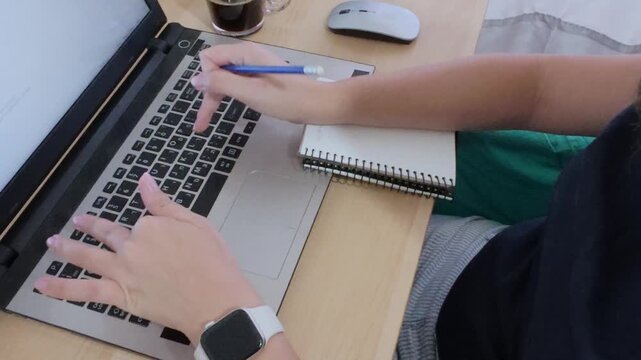 Close-up of hands multitasking, writing in a notebook while using a laptop with Hebrew text.