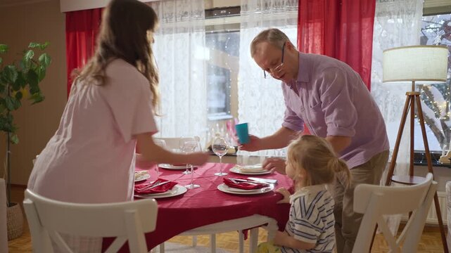 Happy family preparing festive dinner together at home, decorating the table near Christmas tree with glowing lights. Cozy holiday atmosphere, family lifestyle and winter celebration.