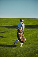 Obraz premium Golfer carrying bag walking across fairway on sunny day. Concept of sports lifestyle, leisure, health, outdoor activity, travel and wellbeing.
