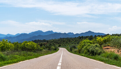 A winding road meanders through the verdant landscape of the Serra de Montsant mountains in Catalonia, Spain. The lush greenery, under a bright sky with scattered clouds.