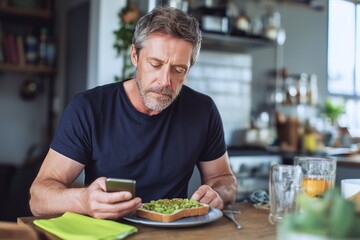 Middle-aged man eating healthy breakfast while scrolling phone. Modern lifestyle, digital habits, and mindful nutrition in cozy home kitchen